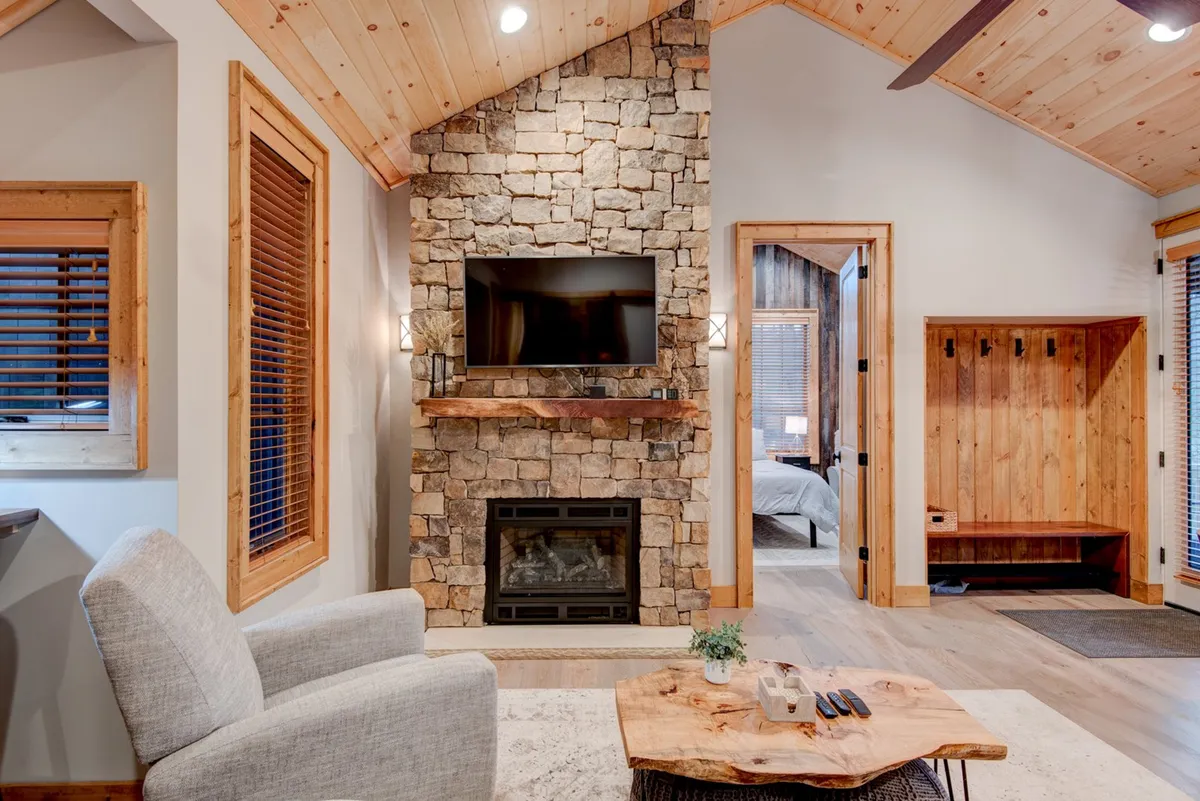 A stone fireplace anchors a cozy living area, accompanied by a comfortable armchair, wooden coffee table, and television. The space is designed for relaxing by the fire, watching TV, or socializing after an active day outdoors.