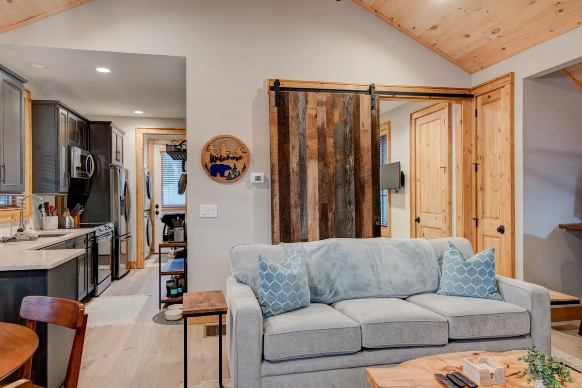 This interior view shows a welcoming living area with a sofa adorned with decorative pillows, a wooden sliding barn door, a welcome sign, and a glimpse into the kitchen. The open layout hints at family gatherings, relaxing moments, and accessible areas for pets to move freely.