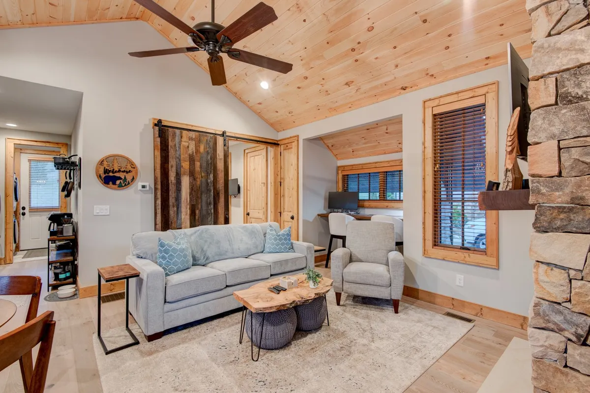 This cozy living room features a comfortable sofa, a rustic coffee table, exposed wooden beam ceilings, and a striking stone fireplace. The arrangement invites relaxation and togetherness, with space for guests to gather around the fireplace or enjoy a quiet moment with a book or cup of coffee.