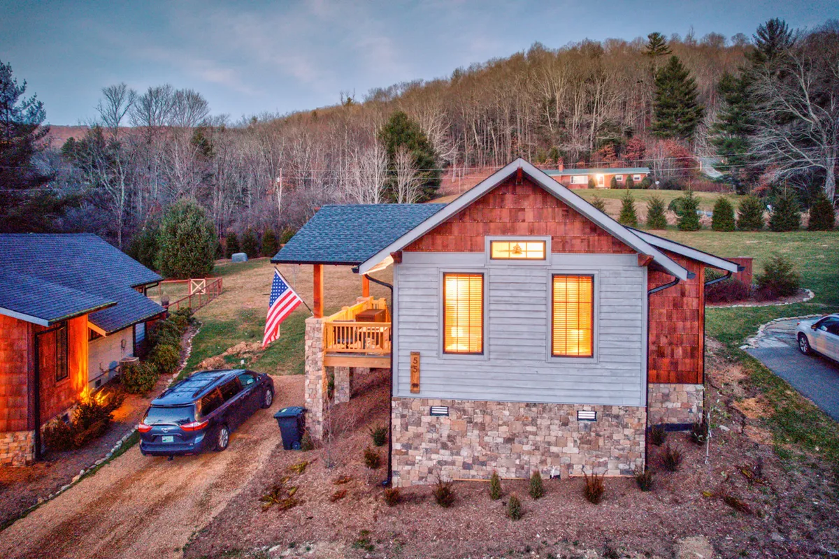 Captured from the front, this photo showcases the home's spacious porch adorned with an American flag, large windows warmly lit from within, and a stone-and-wood facade. The inviting atmosphere hints at a peaceful spot to unwind after a day in town.