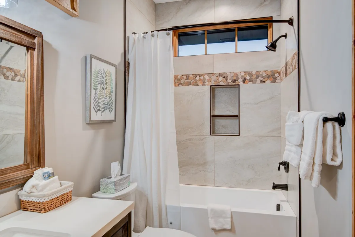 A bright bathroom with a tiled tub/shower combo, a decorative tile stripe, a large mirror, and a bathtub perfect for a relaxing soak. The space is inviting and ideal for self-care routines.