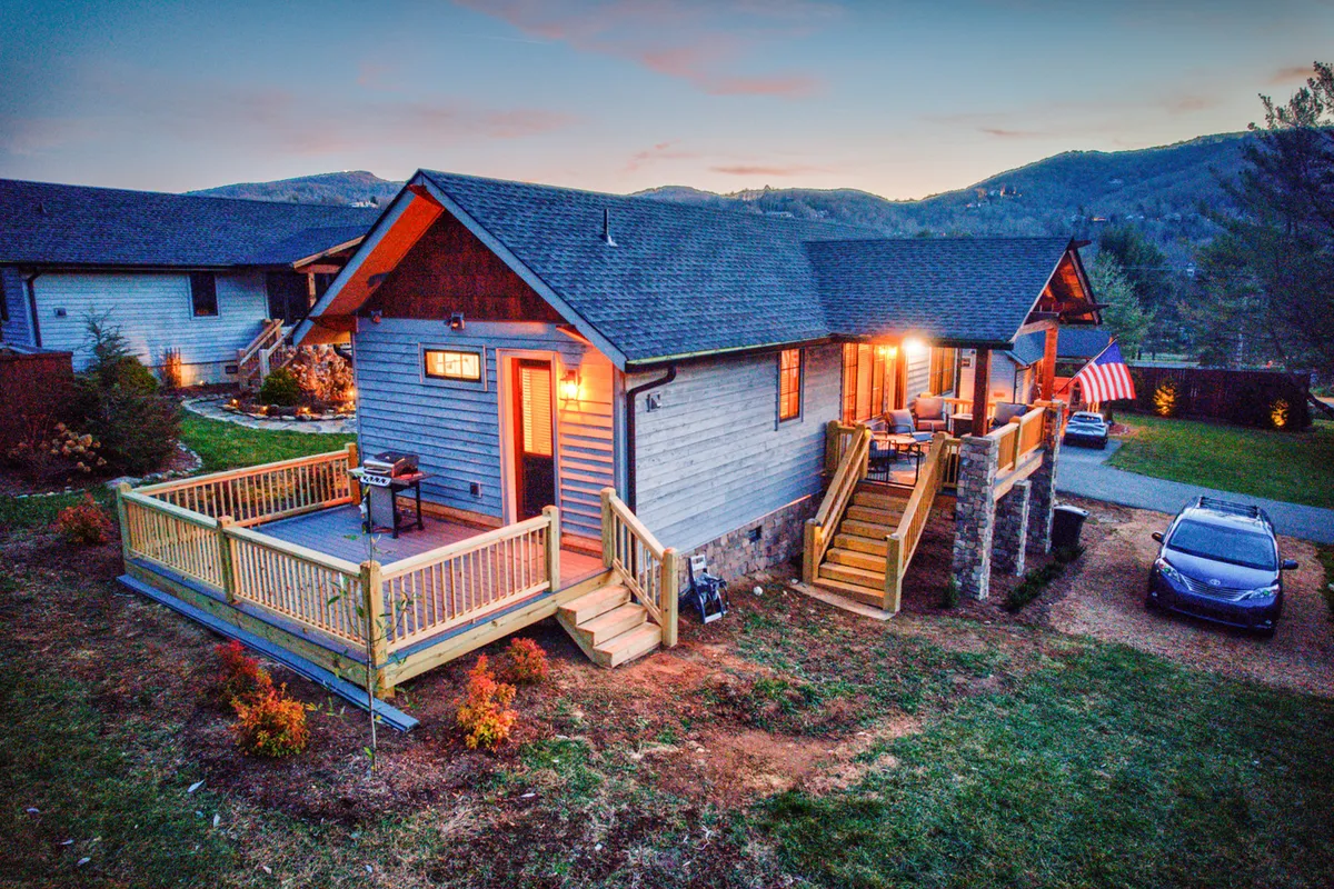 A wooden deck adorned with an American flag overlooks the natural landscape. Illuminated windows hint at warmth inside, while the deck itself offers a comfortable place for outdoor dining, stargazing, or relaxing with friends after mountain adventures.