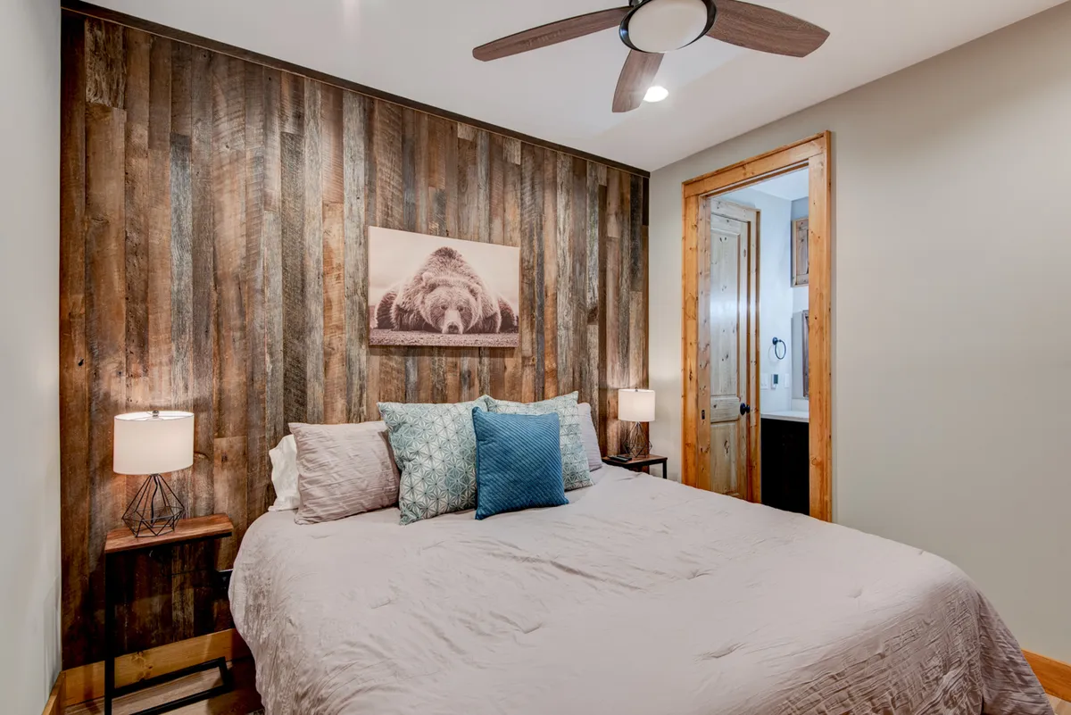 A cozy king bedroom featuring a wooden accent wall, a bed adorned with plush pillows and linens, and a large bear photograph above the headboard. Natural light filters in, creating a restful and inviting atmosphere perfect for relaxation.