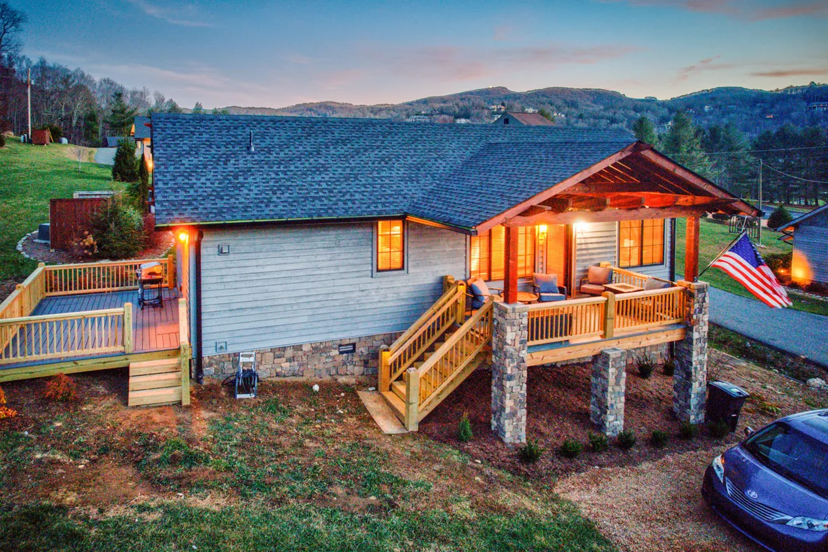 The hero image shows the exterior of a charming single-family home with a welcoming front porch furnished with comfortable seating, supported by sturdy stone pillars. An American flag flutters by the entrance, and the porch is perfectly positioned for relaxing evenings or outdoor gatherings, all set against a serene, mountain-town backdrop.