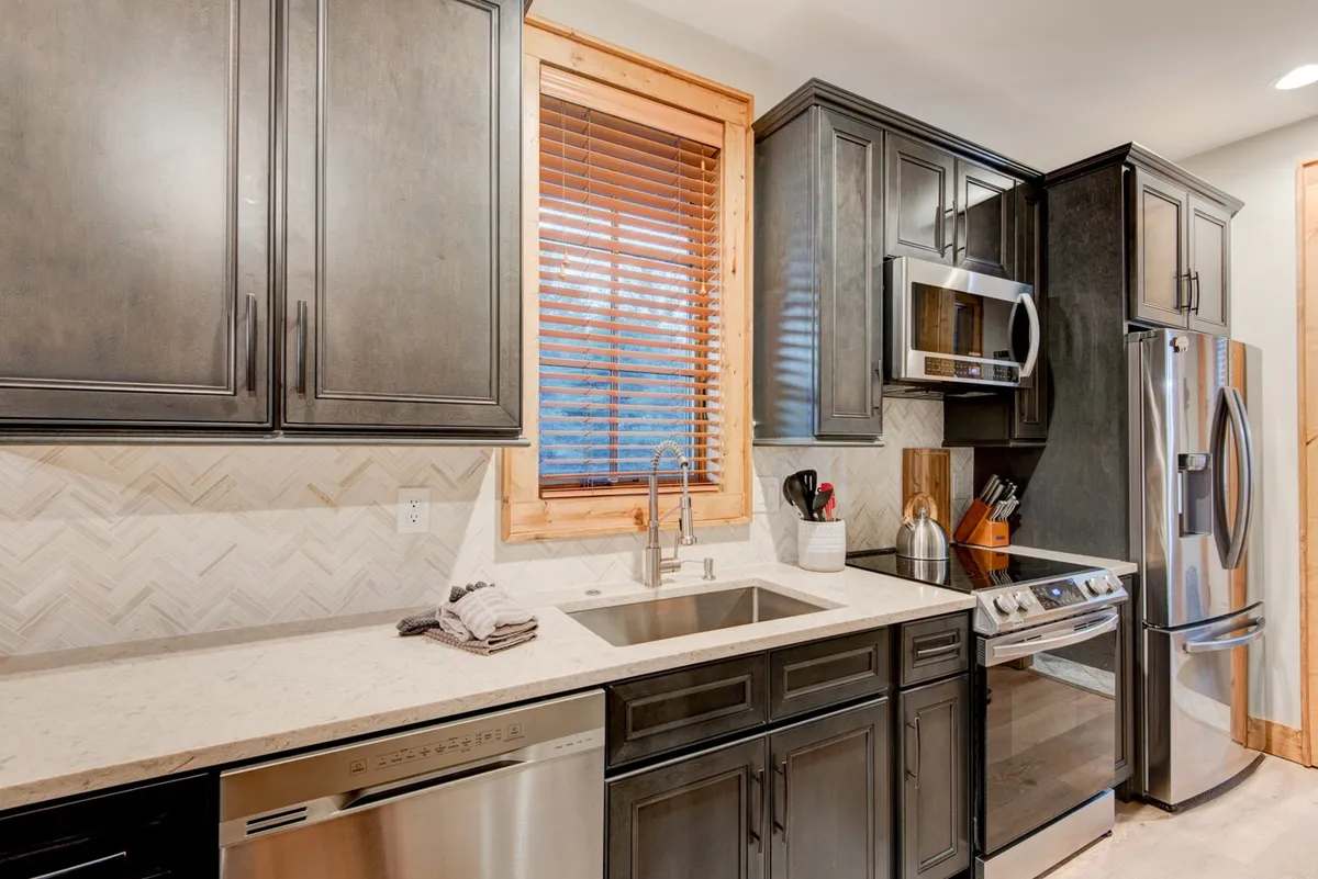 A modern kitchen with stainless steel appliances, a distinctive herringbone tile backsplash, and a warm wooden window frame. The space is designed for both culinary creativity and socializing, as it easily accommodates cooking as a group or entertaining guests.