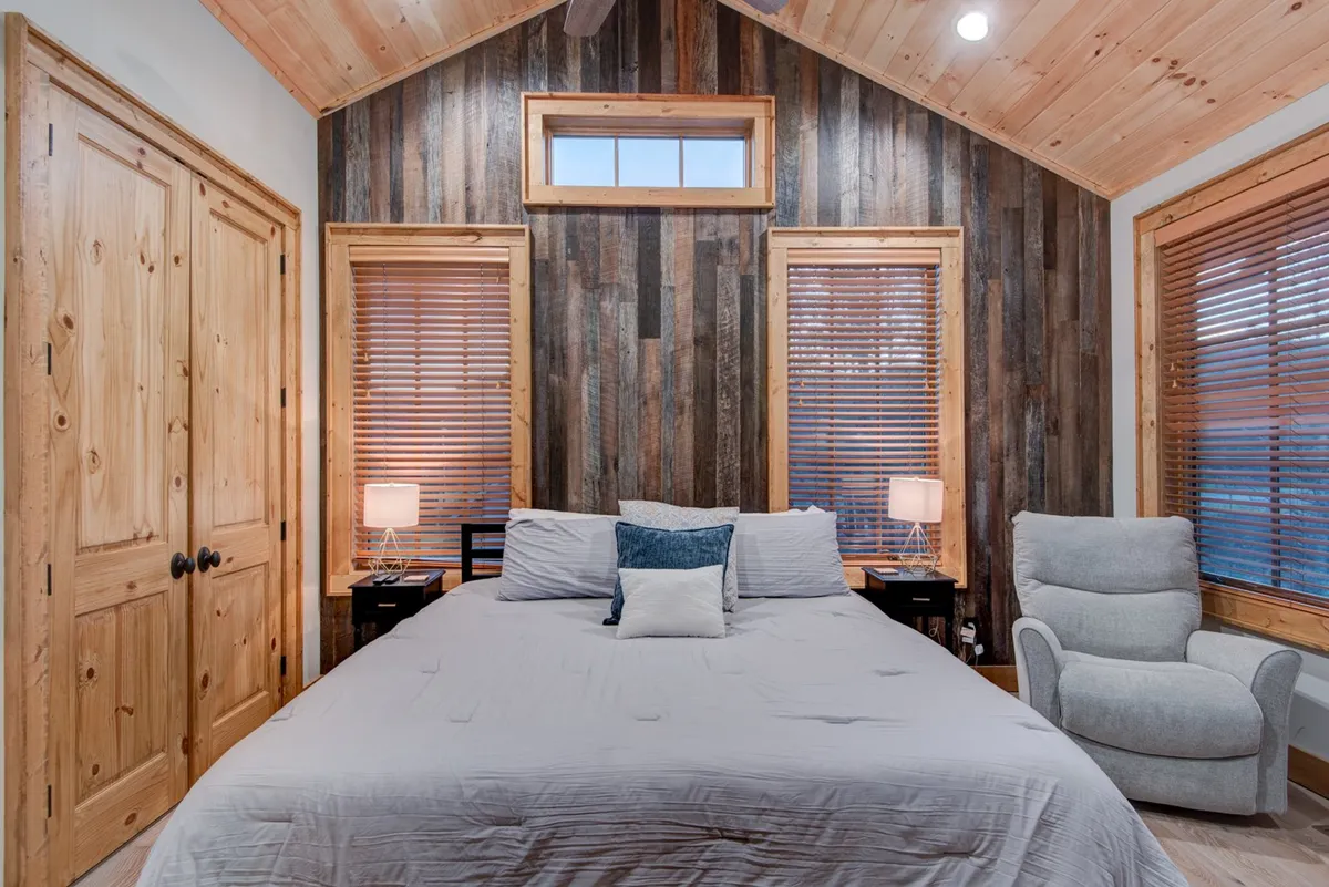 A second king bedroom with a warm wooden feature wall, a bed dressed in neutral bedding, bedside lamps, a recliner chair, and windows with blinds. The soft sunlight creates a welcoming and tranquil setting ideal for reading or unwinding.