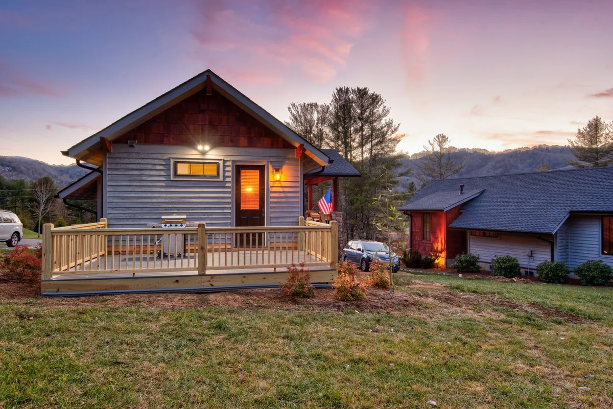 This exterior shot features the house with a welcoming porch overlooking a scenic mountain view during sunset. The American flag waves gently, and the warm glow of the house suggests a cozy, inviting retreat. The porch appears ready for outdoor dining, barbecuing, or relaxing after a stroll downtown.